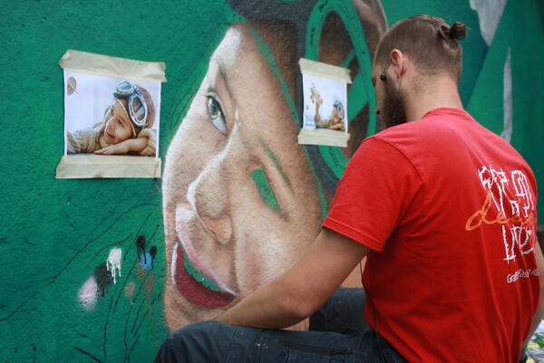 Graffiti-Künstler in rotem T-Shirt bemalt eine grüne Wand mit Hilfe einer Fotovorlage